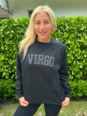 A woman wearing a black sweatshirt with the word 'VIRGO' printed in gray letters, standing in front of a hedge.