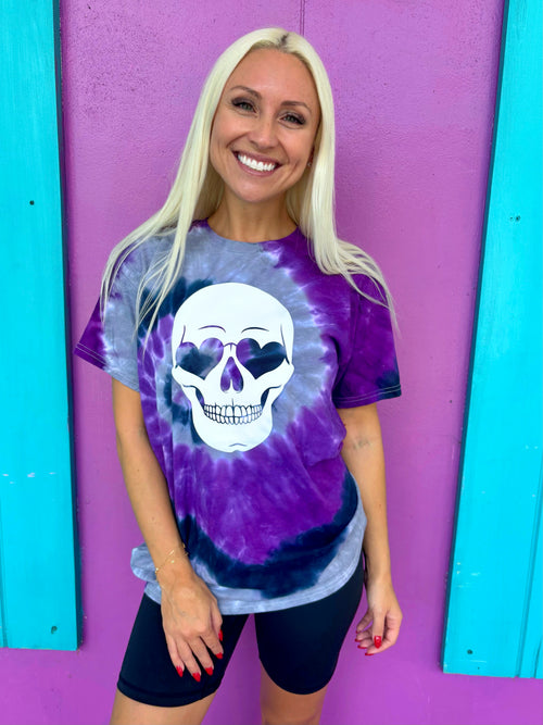 A person wearing a tie-dye t-shirt with a white skull design and purple background, standing in front of a purple wall.