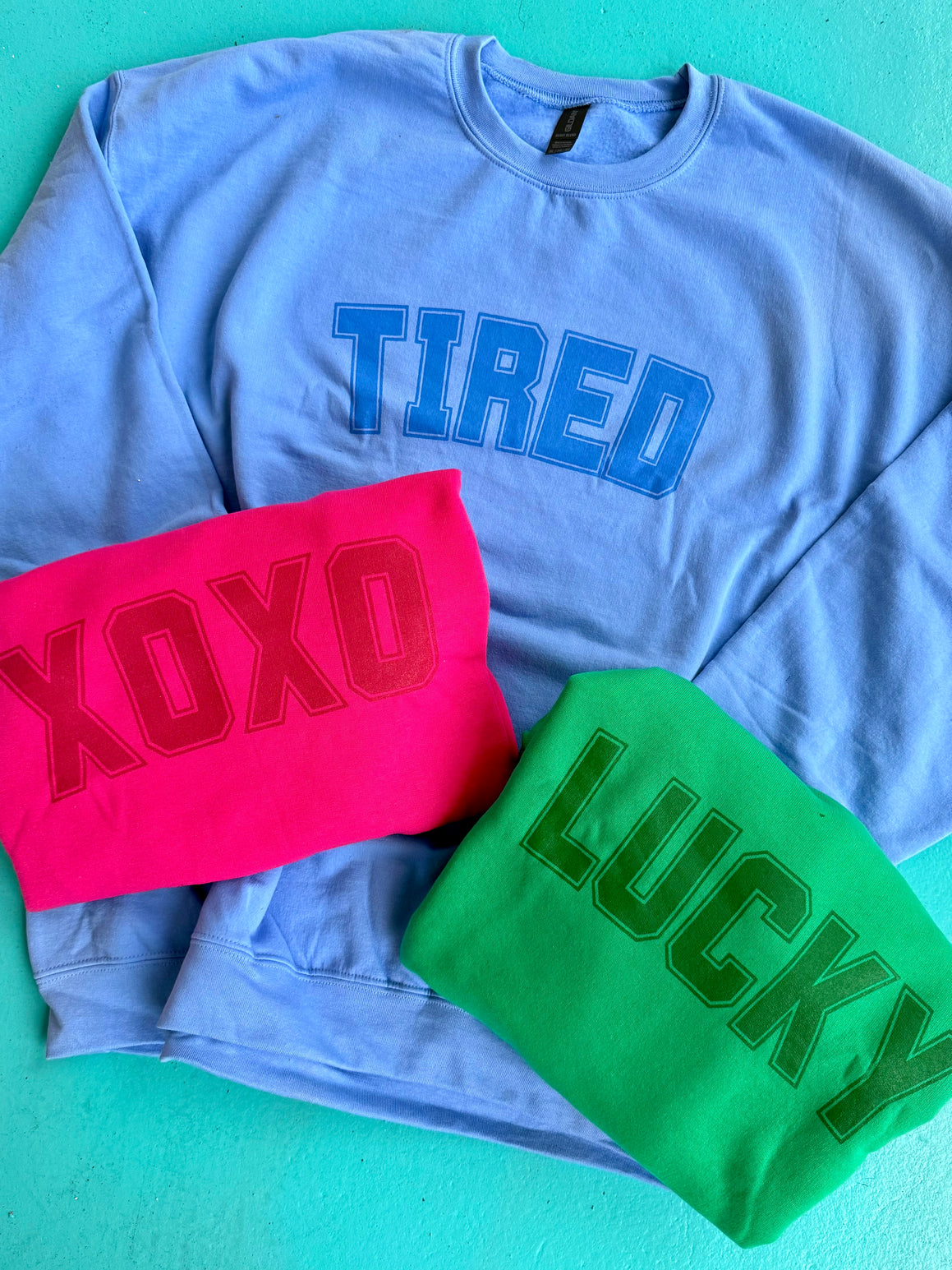 A collection of hoodies displayed, with one in blue featuring the word 'TIRED' in bold letters, another in pink with 'XOXO' and a green hoodie with the word 'LUCKY' in bold letters.