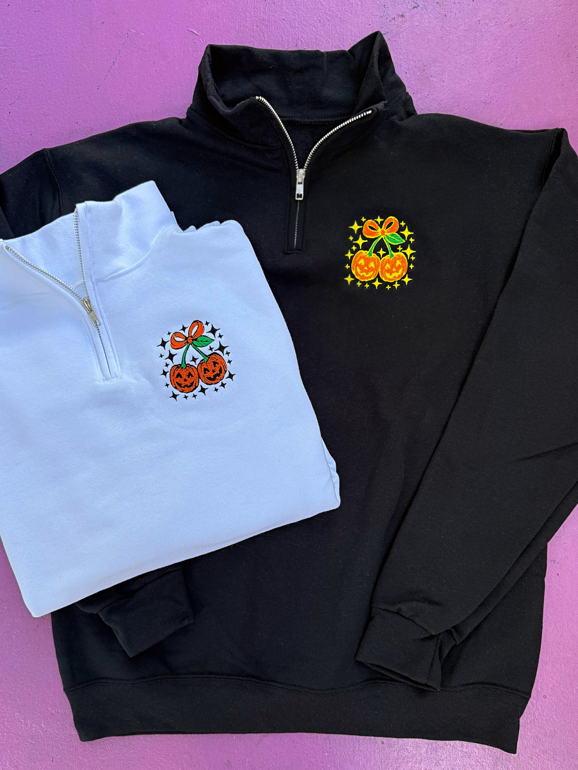 Black and white quarter-zip sweatshirts with Halloween-themed embroidery on a pink background