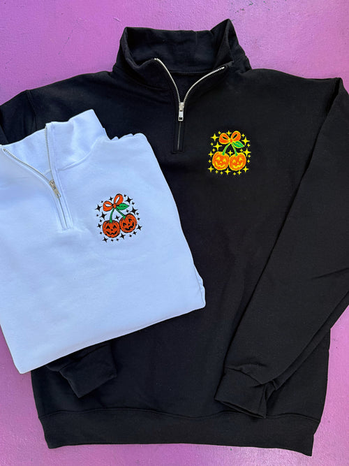Black and white quarter-zip sweatshirts with Halloween-themed embroidery on a pink background