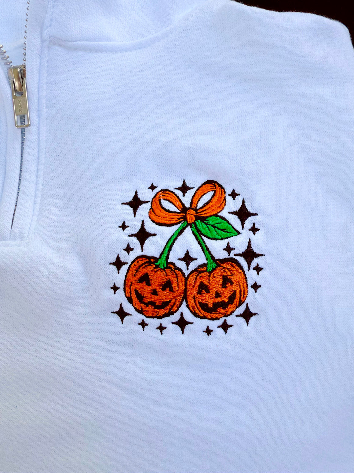 White quarter-zip with Halloween-themed design featuring pumpkins and a bow on a white background.