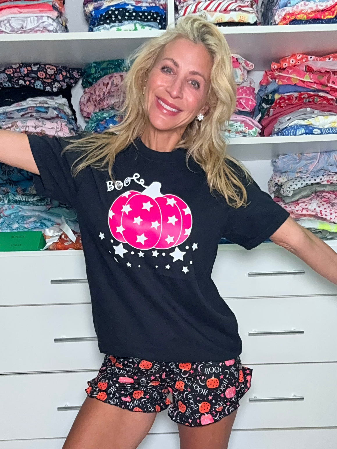 Woman wearing a black t-shirt with a pink pumpkin design and patterned shorts in front of shelves with fabric.