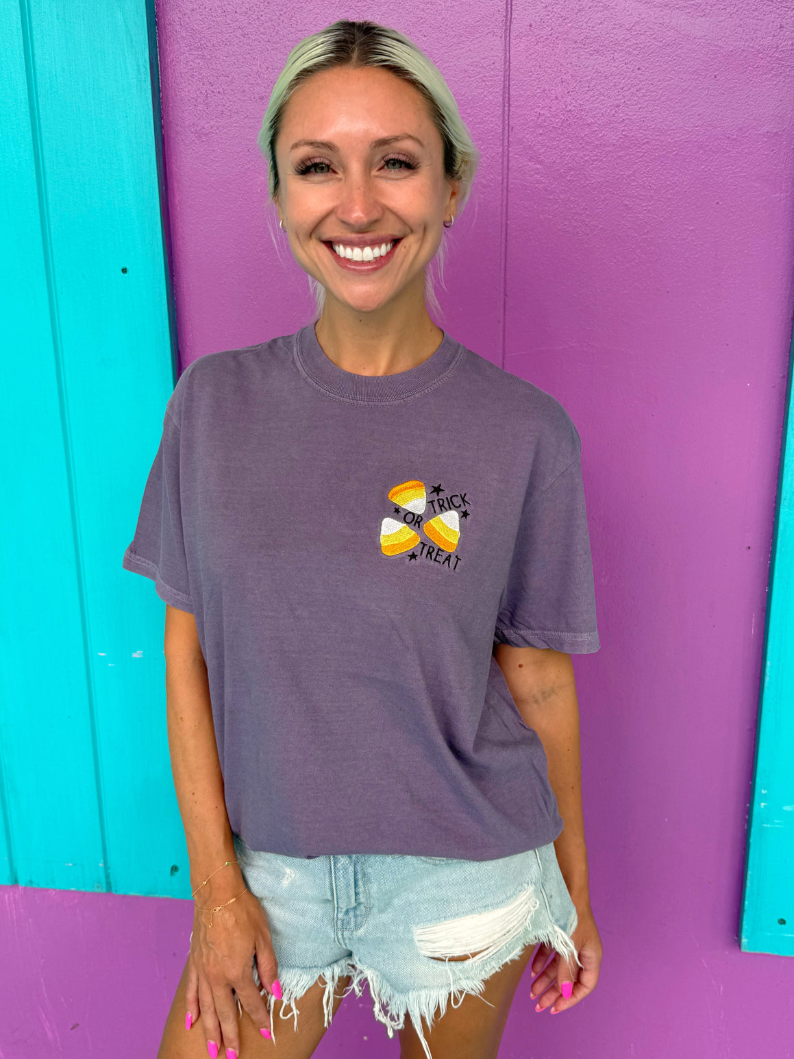 Woman wearing a purple t-shirt with colorful candy corn embroidery against a multicolored wall.