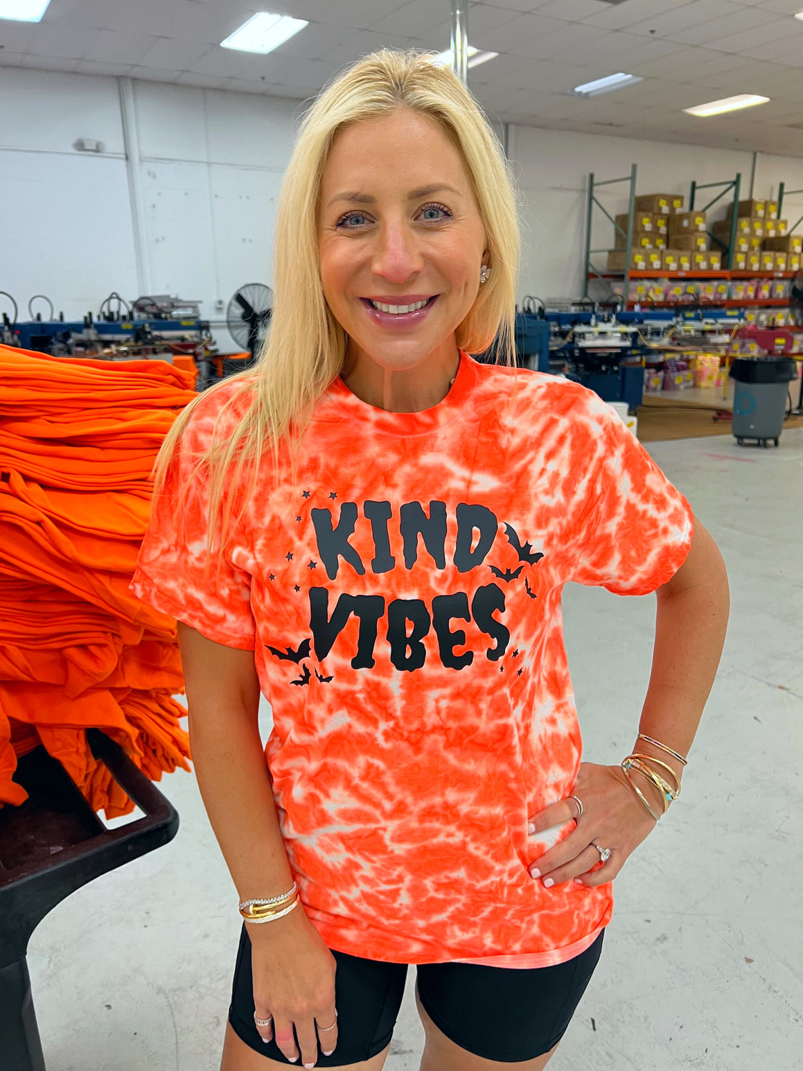 Person wearing an orange tie-dye shirt with 'KIND VIBES' text in a warehouse setting.


