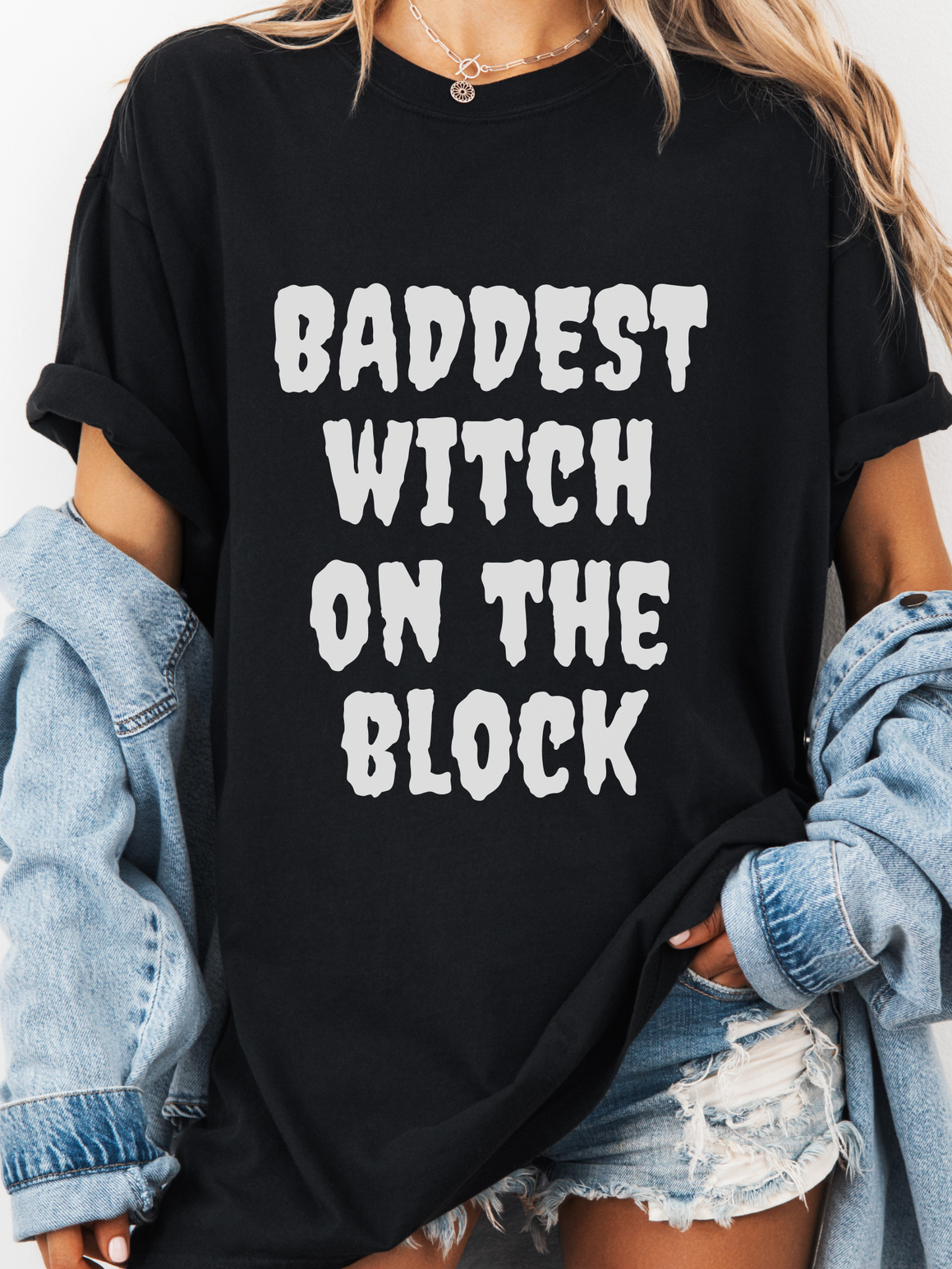 Black t-shirt with 'Baddest Witch on the Block' text worn by a person with denim jacket and shorts.